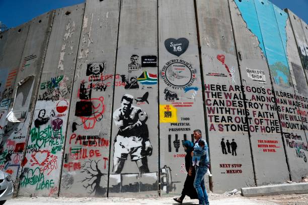 Palestinian family walk past a graffiti painted on the Israeli apartheid wall in the West Bank city of Bethlehem